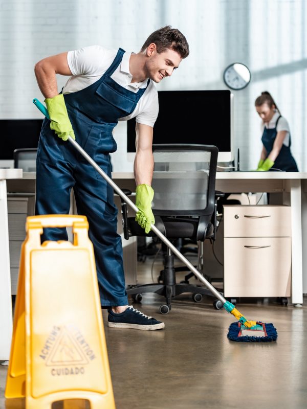 smiling cleaner washing floor with mop near colleague cleaning desk